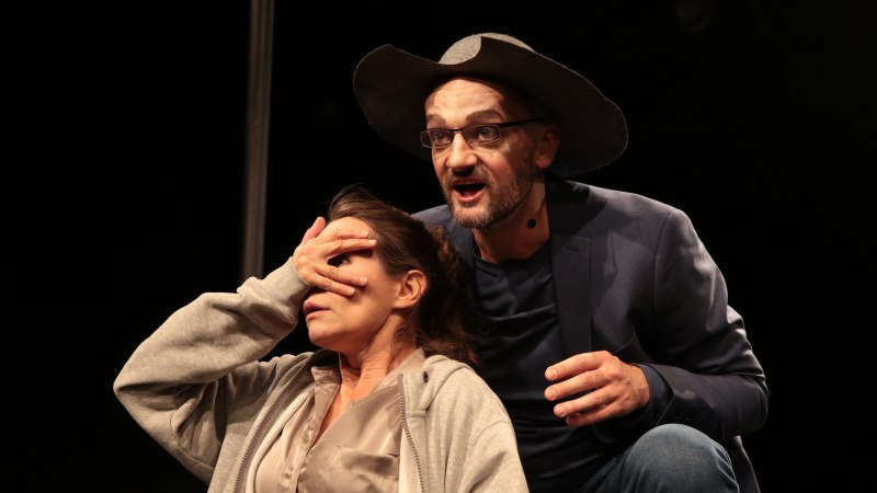Ein Mann mit Hut und Brille spricht auf der Bühne, während er die Augen einer Frau verdeckt. Beide tragen Mikrofone., © Theater tri-bühne, Stefan Kirchknopf