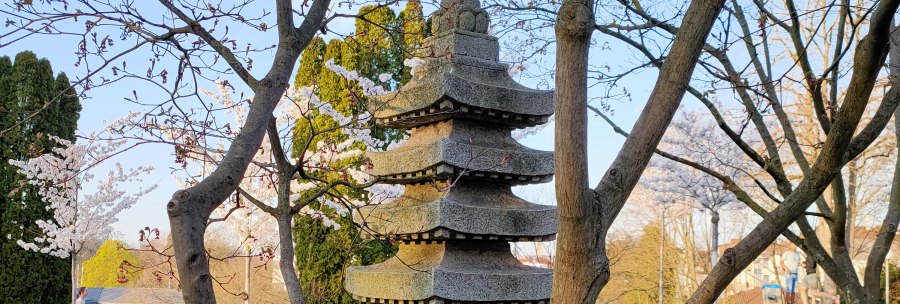 A stone pagoda stands in a garden, surrounded by blossoming trees and green lawns. The sky is clear and blue., © SMG A stone pagoda stands in a garden, surrounded by blossoming trees and green lawns. The sky is clear and blue., © SMG