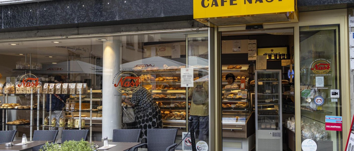 Außenansicht des Cafe Nast mit großen Fenstern, durch die Backwaren und Kunden zu sehen sind. Ein gelbes Schild mit dem Café-Namen hängt über dem Eingang., © SMG, Sarah Schmid