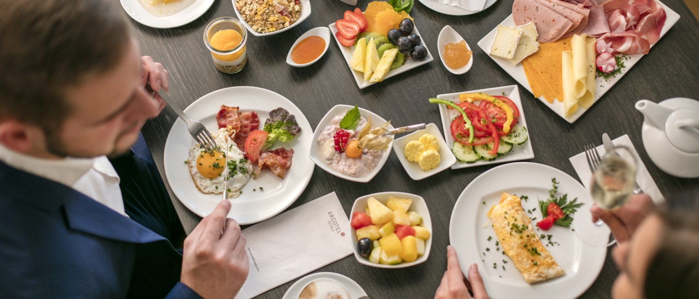 A table with a varied breakfast buffet: eggs, cold cuts, fruit, muesli and drinks. Two people sit at the table and eat., © ARCOTEL Hotels A table with a varied breakfast buffet: eggs, cold cuts, fruit, muesli and drinks. Two people sit at the table and eat., © ARCOTEL Hotels