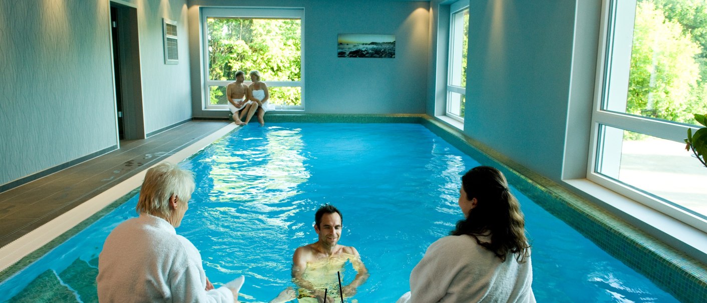 An indoor pool with several people in bathrobes and towels. Some sit at the edge of the pool while one person stands in the water., © Hotel Fortuna