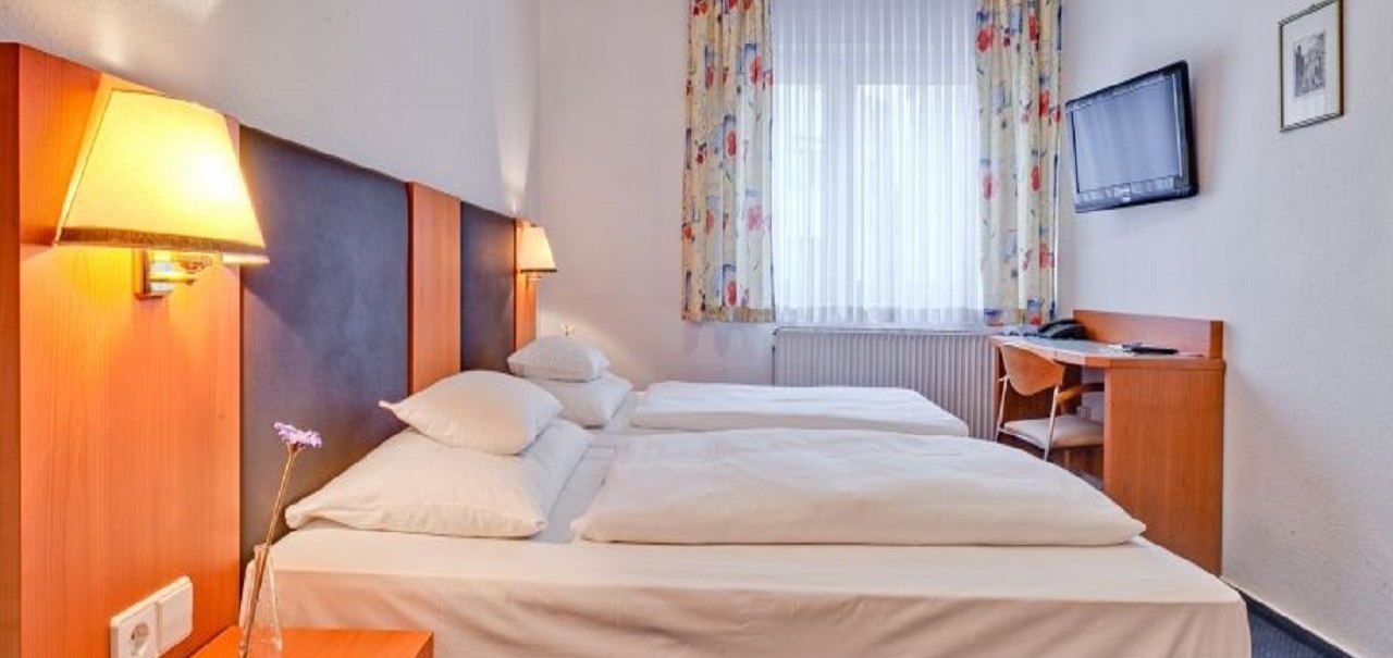 Ein gemütliches Doppelzimmer mit zwei Betten, einem Schreibtisch, einem Stuhl und einem Wandfernseher. Die Vorhänge sind bunt gemustert., © ARNOTEL GmbH