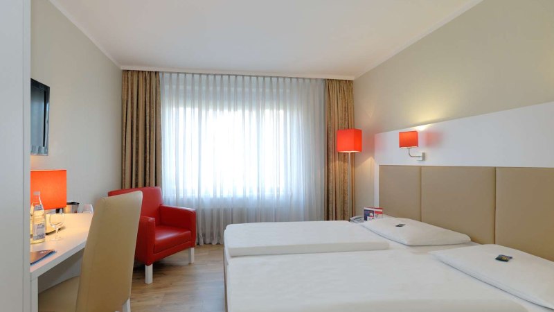 Modernes Hotelzimmer mit zwei Einzelbetten, rotem Sessel, Schreibtisch und Fenster mit Vorhängen. Rote Lampen sorgen für Akzente., © Hotel Stuttgart-Sindelfingen City by Tulip Inn Modernes Hotelzimmer mit zwei Einzelbetten, rotem Sessel, Schreibtisch und Fenster mit Vorhängen. Rote Lampen sorgen für Akzente., © Hotel Stuttgart-Sindelfingen City by Tulip Inn