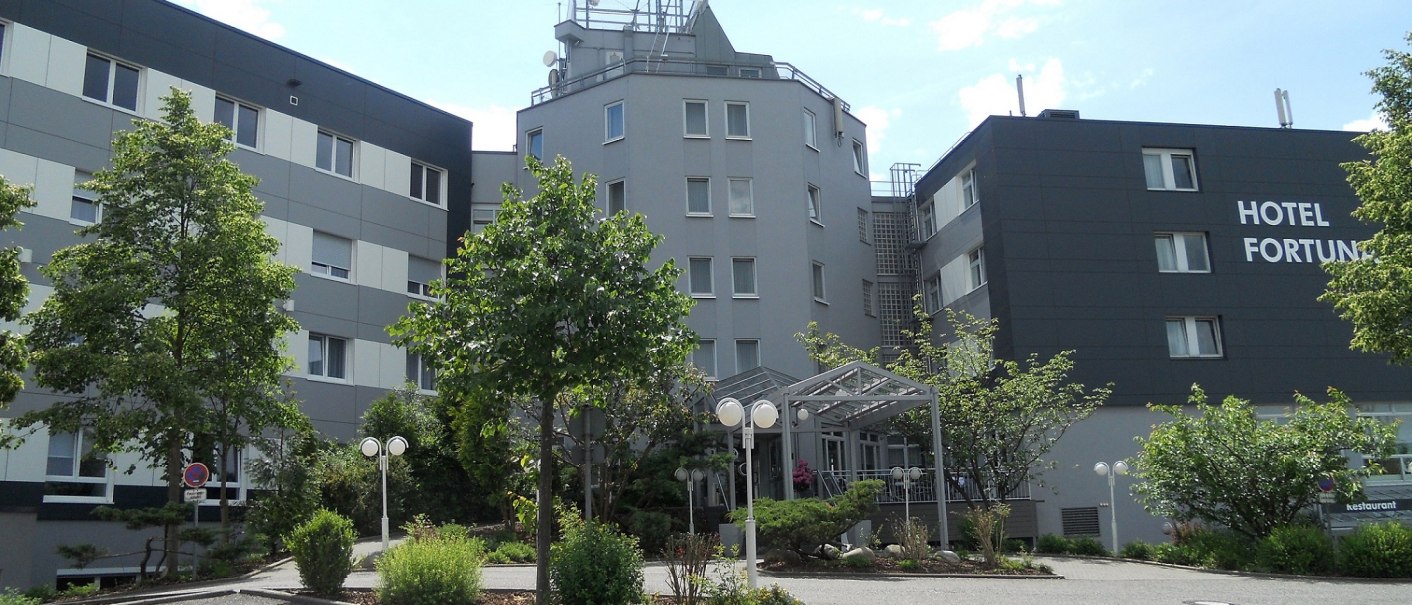 Modernes Hotel mit grauer Fassade, umgeben von Bäumen und Pflanzen. Der Himmel ist klar und sonnig., © Hotel Fortuna