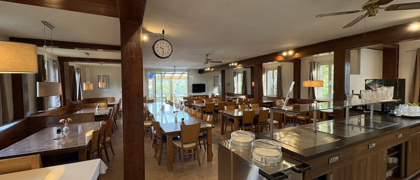 Ein gemütliches Restaurant mit Holzmöbeln, gedeckten Tischen und einer großen Uhr an der Decke. Große Fenster lassen viel Licht herein., © TOMAS Ein gemütliches Restaurant mit Holzmöbeln, gedeckten Tischen und einer großen Uhr an der Decke. Große Fenster lassen viel Licht herein., © TOMAS