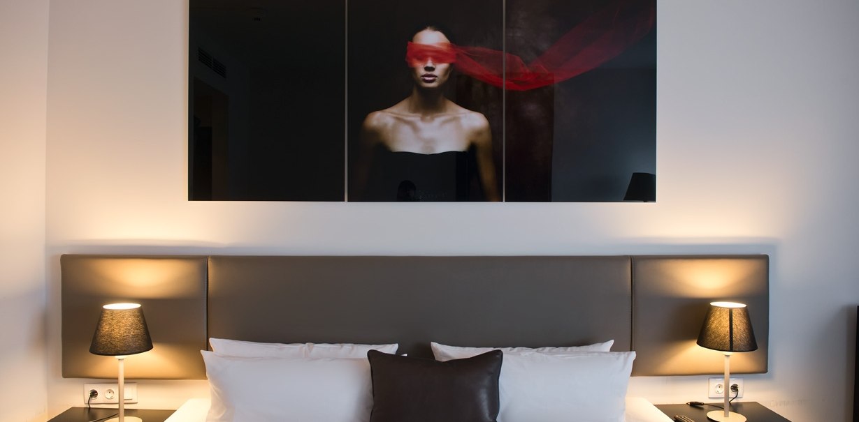 Modern hotel room with double bed, two bedside lamps and a work of art above the bed showing a person wearing a red scarf., © Wyndham Stuttgart Airport Messe Modern hotel room with double bed, two bedside lamps and a work of art above the bed showing a person wearing a red scarf., © Wyndham Stuttgart Airport Messe