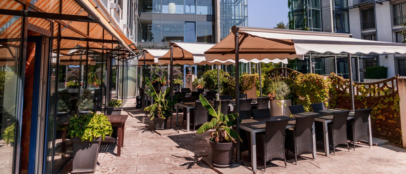 Sunny outdoor area of a café with tables, chairs and plants under awnings. Modern glass façade in the background., © PLAZA Hotelgroup GmbH