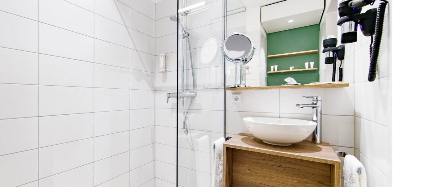 Modern bathroom with glass shower, round washbasin on wooden shelf, mirror and hairdryer on the wall. White tiles and green accent on the wall., © Arthotel ANA Momentum Modern bathroom with glass shower, round washbasin on wooden shelf, mirror and hairdryer on the wall. White tiles and green accent on the wall., © Arthotel ANA Momentum