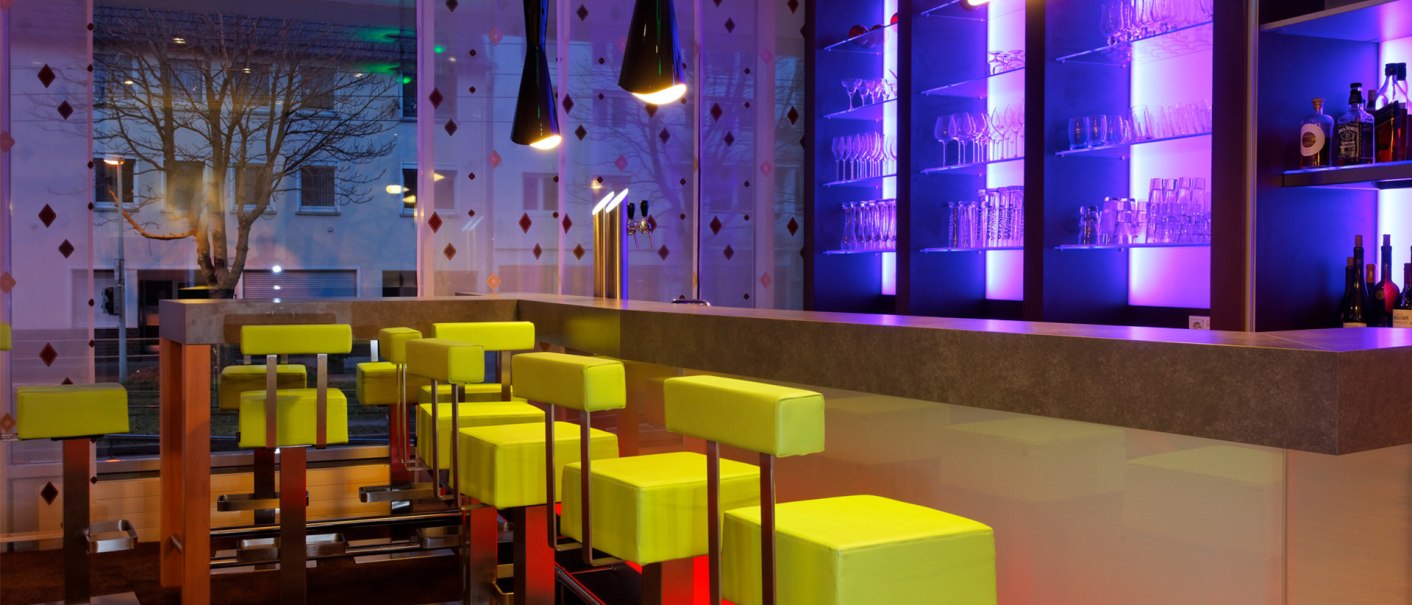 Moderne Bar mit leuchtend grünen Hockern, farbiger Beleuchtung und Glasregalen. Große Fensterfront mit Baum im Hintergrund., © Ibis Styles Stuttgart Moderne Bar mit leuchtend grünen Hockern, farbiger Beleuchtung und Glasregalen. Große Fensterfront mit Baum im Hintergrund., © Ibis Styles Stuttgart
