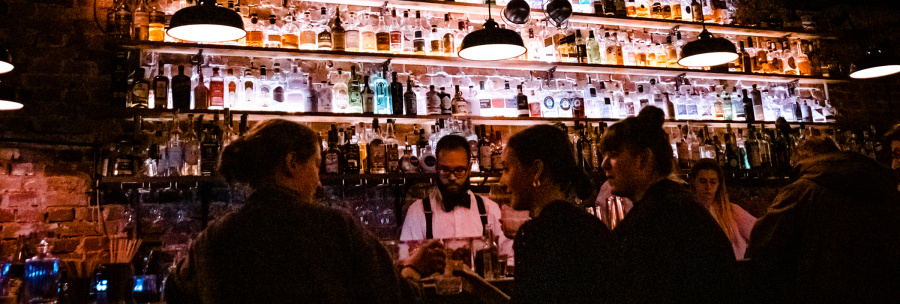 Eine stimmungsvolle Bar mit beleuchteten Flaschenregalen. Menschen unterhalten sich, w&auml;hrend ein Barkeeper Getr&auml;nke zubereitet., &copy; Stuttgart-Marketing GmbH, Sarah Schmid