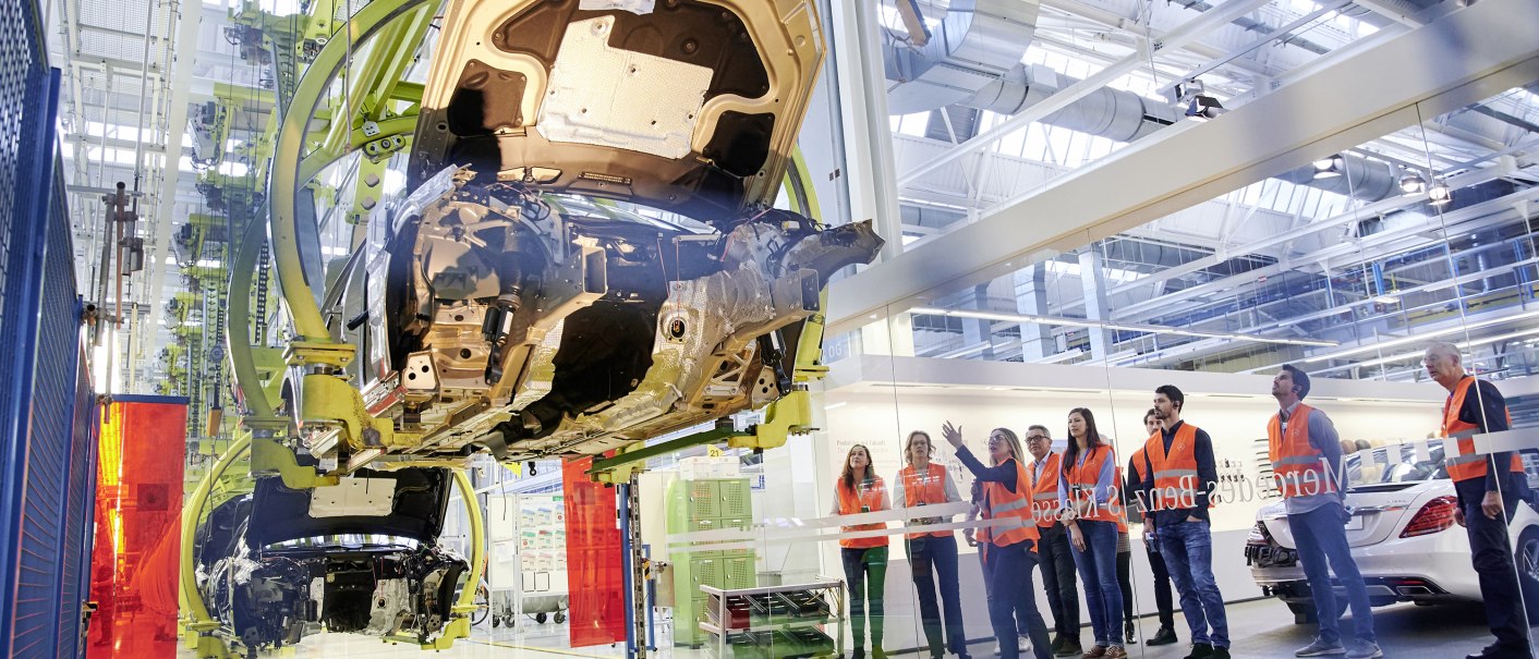 Visitors in orange vests observe car production at the Mercedes-Benz Customer Center. A vehicle chassis hangs on a conveyor belt., © Mercedes-Benz Kundencenter