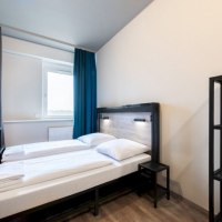 Modernes Doppelzimmer mit zwei Einzelbetten, einem kleinen Nachttisch und einem Regal. Helle Wände und Vorhänge in Blau und Grau., © A&O Hostels Marketing GmbH Modernes Doppelzimmer mit zwei Einzelbetten, einem kleinen Nachttisch und einem Regal. Helle Wände und Vorhänge in Blau und Grau., © A&O Hostels Marketing GmbH