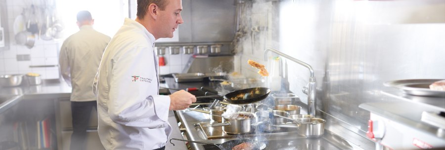 A chef in a white uniform fries meat in a pan in a professional kitchen. Another chef can be seen in the background., © SMG, Jean-Claude Winkler