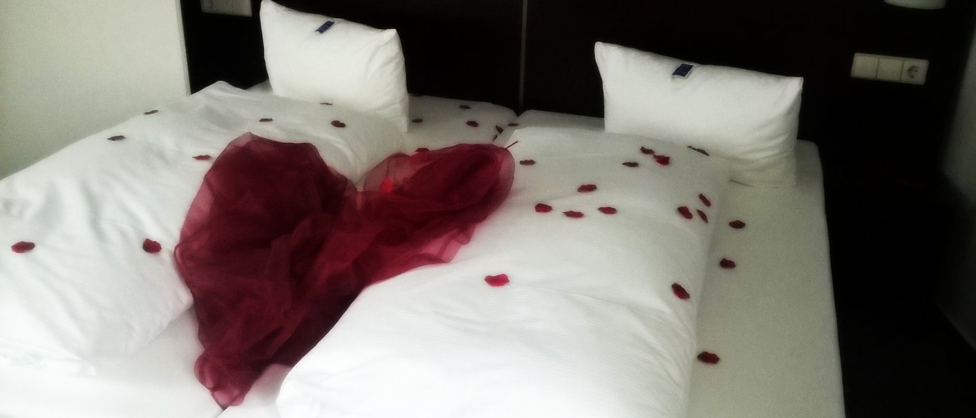 Ein Hotelbett mit weißen Kissen, dekoriert mit roten Rosenblättern und einem roten Tuch in Herzform., © AKZENT Hotel Torgauer Hof.