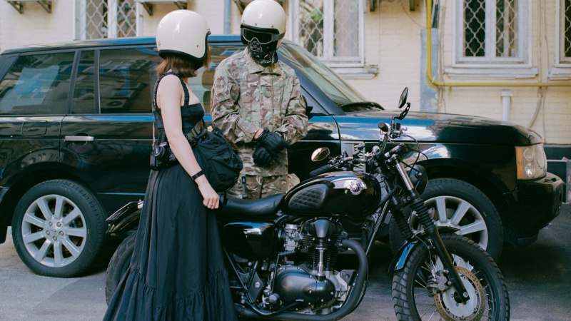 Zwei Personen mit Helmen stehen neben einem Motorrad vor einem schwarzen Auto. Eine Person trägt ein schwarzes Kleid, die andere Tarnkleidung., © Anya Tsaruk Zwei Personen mit Helmen stehen neben einem Motorrad vor einem schwarzen Auto. Eine Person trägt ein schwarzes Kleid, die andere Tarnkleidung., © Anya Tsaruk