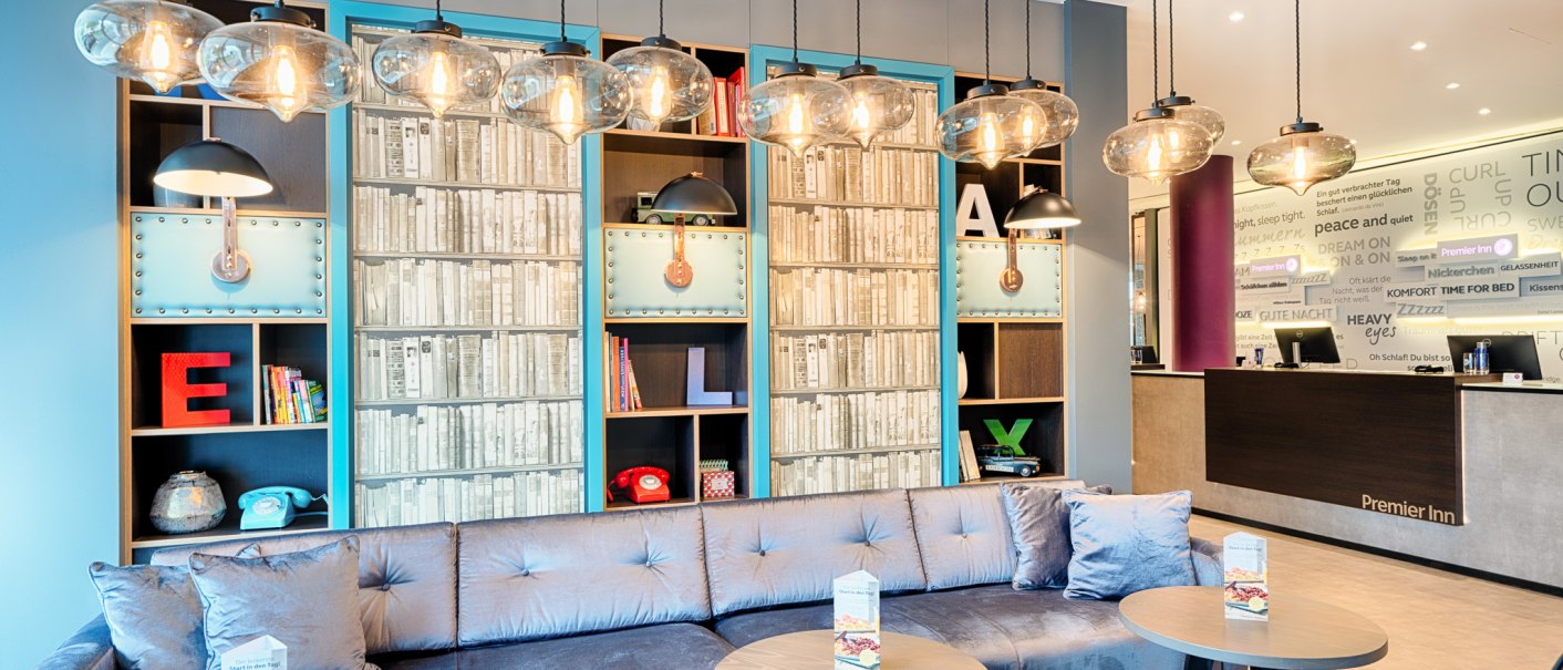 Modern lobby with blue couch, round tables and decorative shelves. Hanging lamps and a reception desk in the background., © Premier Inn Germany Modern lobby with blue couch, round tables and decorative shelves. Hanging lamps and a reception desk in the background., © Premier Inn Germany