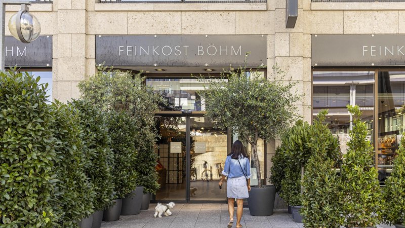 Eingang von Feinkost Böhm, umgeben von grünen Pflanzen. Eine Frau mit einem kleinen weißen Hund betritt das Geschäft., © SMG, Sarah Schmid Eingang von Feinkost Böhm, umgeben von grünen Pflanzen. Eine Frau mit einem kleinen weißen Hund betritt das Geschäft., © SMG, Sarah Schmid