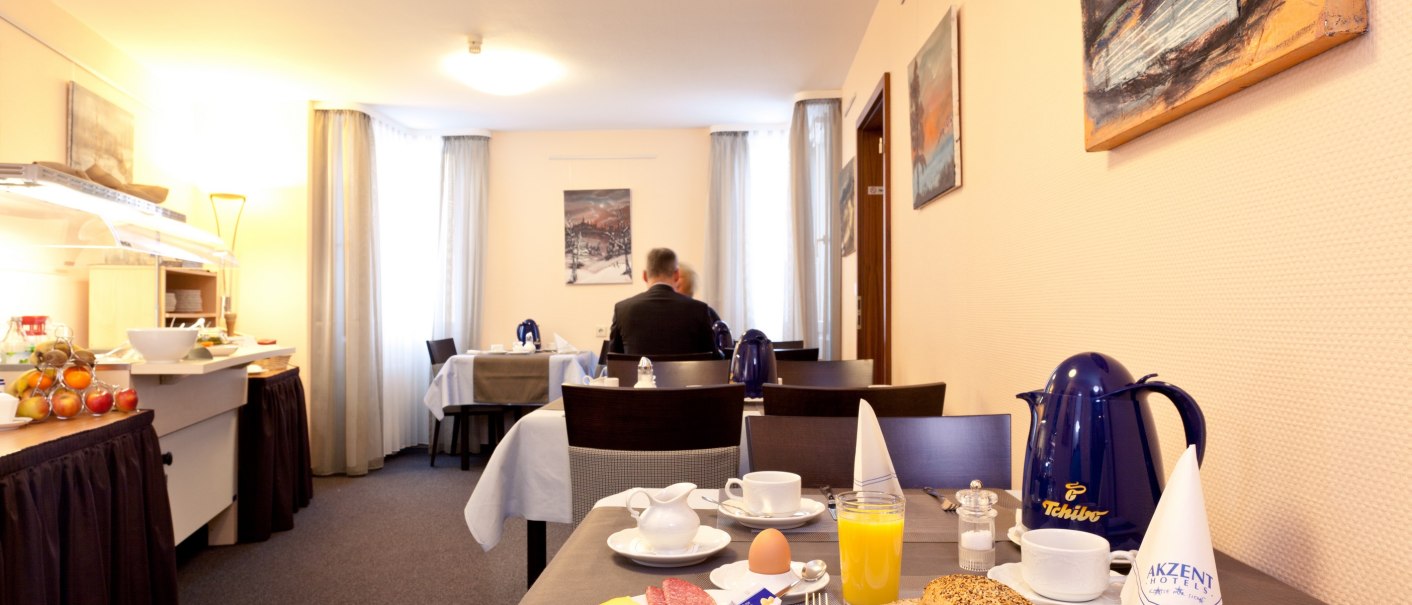 Frühstücksraum im AKZENT Hotel Torgauer Hof mit gedecktem Tisch, Frühstücksbuffet und Kunst an den Wänden. Eine Person sitzt im Hintergrund., © AKZENT Hotel Torgauer Hof