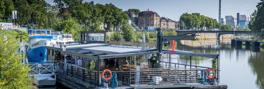 Ein schwimmendes Restaurant auf dem Neckar, umgeben von gr&uuml;ner Vegetation und st&auml;dtischen Geb&auml;uden im Hintergrund., &copy; all copyrights are reserved by maks richter
