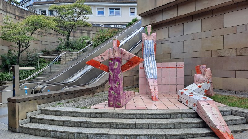 Bunte Skulpturen von Wolfgang Thiel stehen vor einer Rolltreppe in einer städtischen Umgebung mit Bäumen und Gebäuden im Hintergrund., © Foto: Wolfgang Thiel Bunte Skulpturen von Wolfgang Thiel stehen vor einer Rolltreppe in einer städtischen Umgebung mit Bäumen und Gebäuden im Hintergrund., © Foto: Wolfgang Thiel