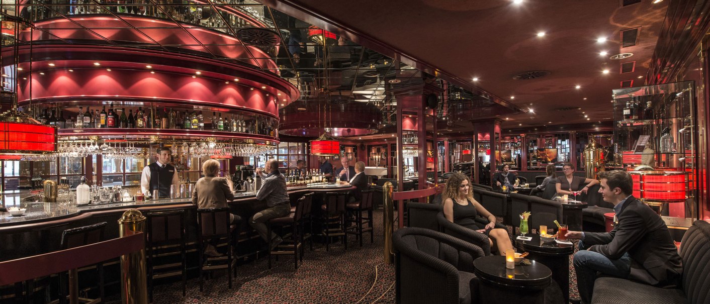 Elegante Bar mit rotem Dekor, Spiegeln und ged&auml;mpfter Beleuchtung. G&auml;ste sitzen an der Theke und an Tischen, w&auml;hrend ein Barkeeper Getr&auml;nke serviert., &copy; Maritim Hotels