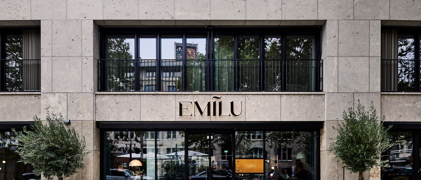 Eingang des EmiLu Hotels mit gro&szlig;en Glasfenstern, Pflanzen in schwarzen T&ouml;pfen und dem Hotelnamen &uuml;ber der T&uuml;r., &copy; EmiLu Hotel GmbH