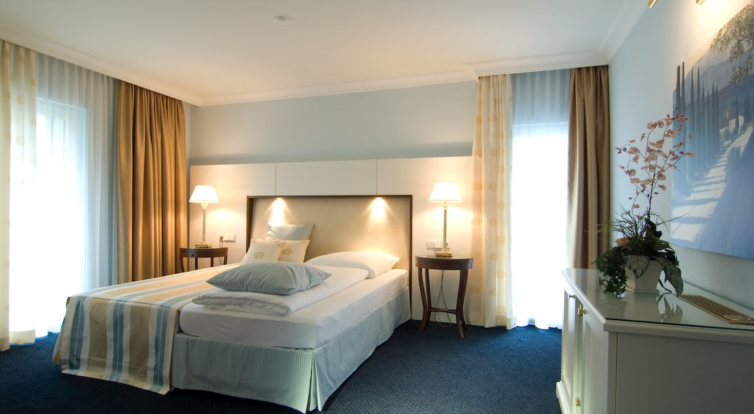 Elegantes Hotelzimmer mit Doppelbett, Nachttischen und Lampen. Helle Vorhänge und ein Bild an der Wand schaffen eine gemütliche Atmosphäre., © Jeanette Bender