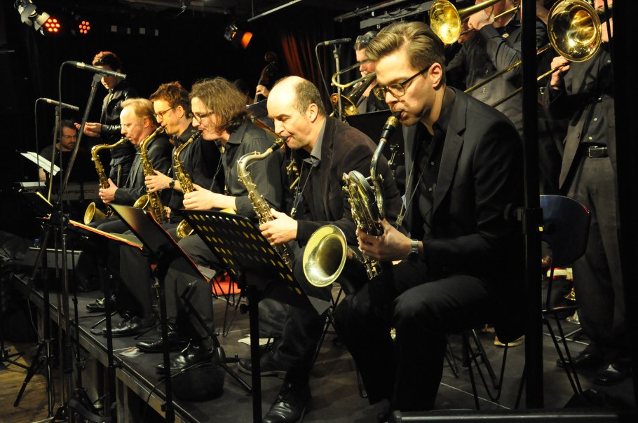 A big band on stage, musicians play saxophones and trombones. The stage is bathed in warm light, music stands are visible., © Pressefoto A big band on stage, musicians play saxophones and trombones. The stage is bathed in warm light, music stands are visible., © Pressefoto
