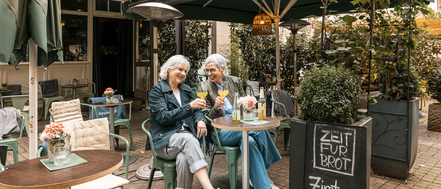 Two women sit in a cozy outdoor café, toasting with drinks and laughing. A sign offers cinnamon buns., © Stuttgart Marketing GmbH - Sarah Schmid