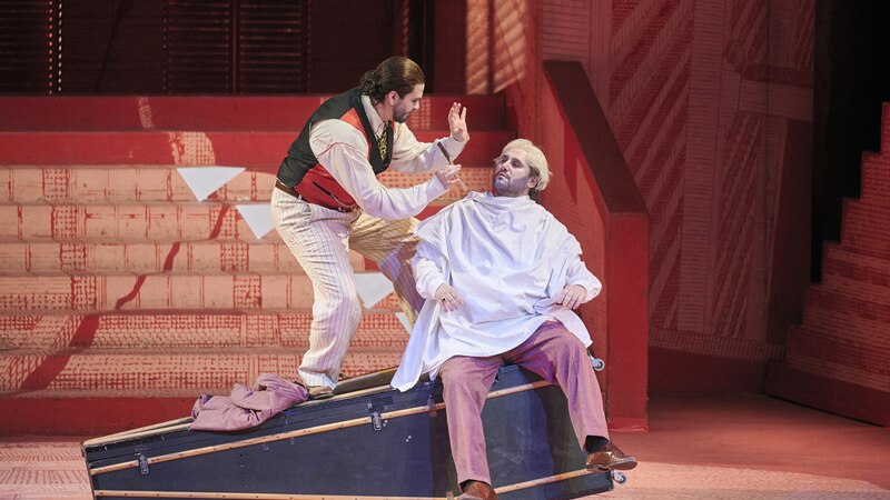 Zwei Darsteller auf einer Opernbühne: Einer als Friseur, der andere im Friseurumhang, auf einer schrägen Plattform sitzend., © Württembergische Staatstheater Stuttgart Zwei Darsteller auf einer Opernbühne: Einer als Friseur, der andere im Friseurumhang, auf einer schrägen Plattform sitzend., © Württembergische Staatstheater Stuttgart
