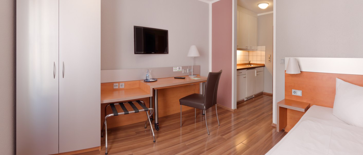 Modernes Einzelzimmer mit Bett, Schreibtisch, Stuhl, Fernseher und Kleiderschrank. Helle Holzmöbel und Laminatboden, kleine Küchenzeile im Hintergrund., © attimo Hotel Stuttgart Modernes Einzelzimmer mit Bett, Schreibtisch, Stuhl, Fernseher und Kleiderschrank. Helle Holzmöbel und Laminatboden, kleine Küchenzeile im Hintergrund., © attimo Hotel Stuttgart