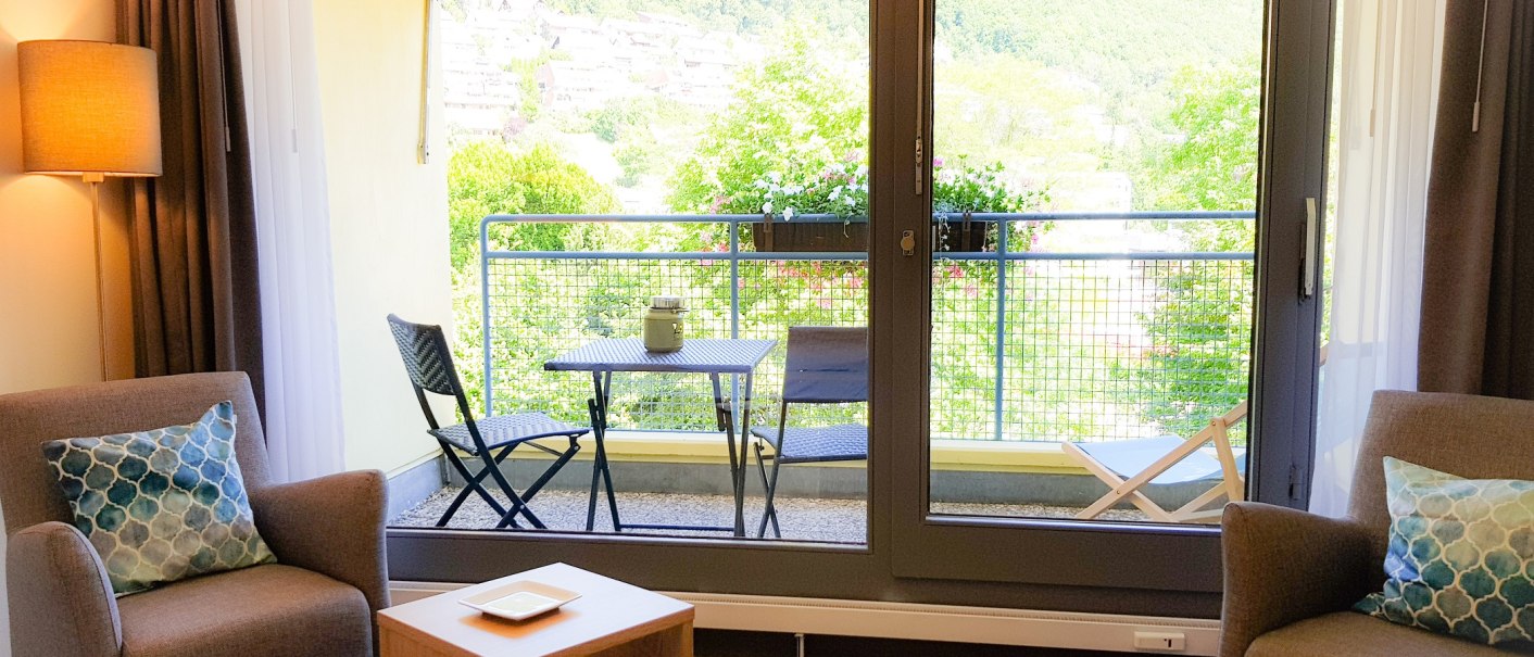 Gemütliches Zimmer mit zwei Sesseln, kleinem Tisch und Balkon mit Sitzgelegenheit. Blick auf grüne Landschaft durch große Fensterfront., © ©hotel-graf-eberhard.de