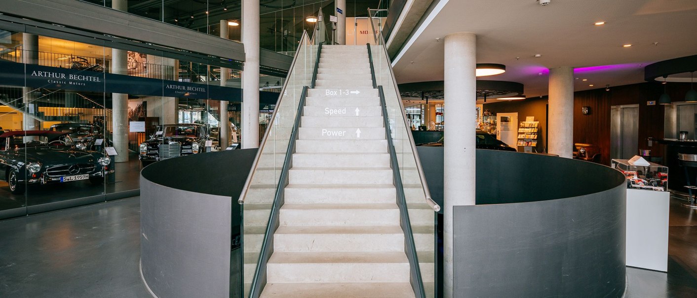 Innenansicht eines Autohauses mit einer Treppe in der Mitte, umgeben von Oldtimern hinter Glas. Moderne Architektur mit Beton und Glas., © V8 HOTEL
