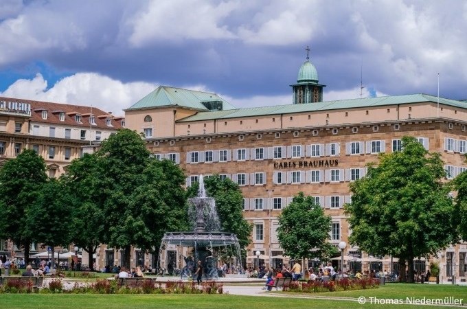 Ein belebter Platz in Stuttgart mit einem Brunnen und dem Gebäude "Carls Brauhaus" im Hintergrund, umgeben von Bäumen und Menschen., © Stuttgart Marketing GmbH