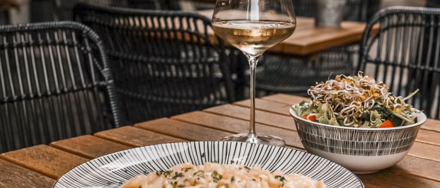 Ein Glas Weißwein, ein Teller Pasta und ein Salat auf einem Holztisch in einem Restaurant mit geflochtenen Stühlen., © SMG, itchyfeet-travel.de