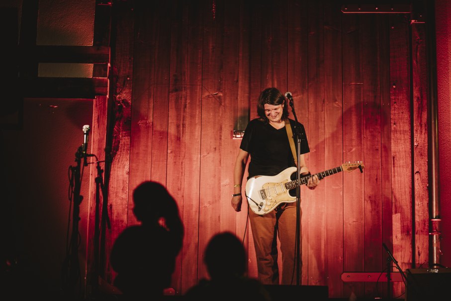 Ein Musiker steht l&auml;chelnd mit einer E-Gitarre auf einer B&uuml;hne vor einer Holzwand, beleuchtet von warmem, rotem Licht., &copy; Stefan Ebert