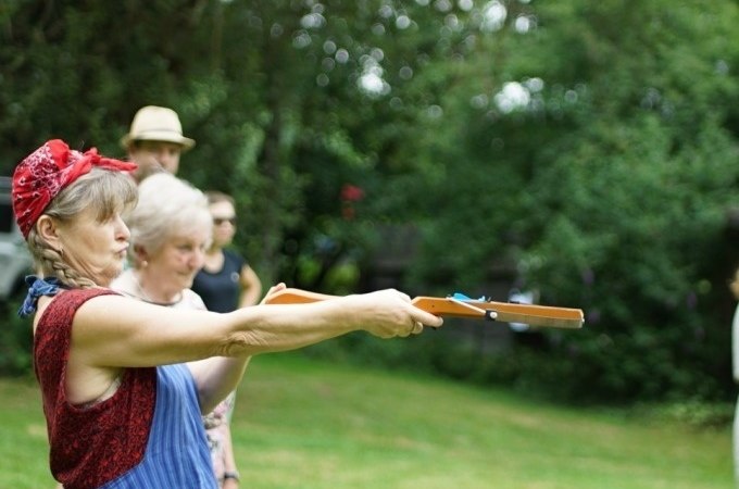 Eine Frau zielt mit einem Spielzeuggewehr bei einem Outdoor-Event. Weitere Personen stehen im Hintergrund auf einer Wiese., © Cool-Tours StattReisen Eine Frau zielt mit einem Spielzeuggewehr bei einem Outdoor-Event. Weitere Personen stehen im Hintergrund auf einer Wiese., © Cool-Tours StattReisen