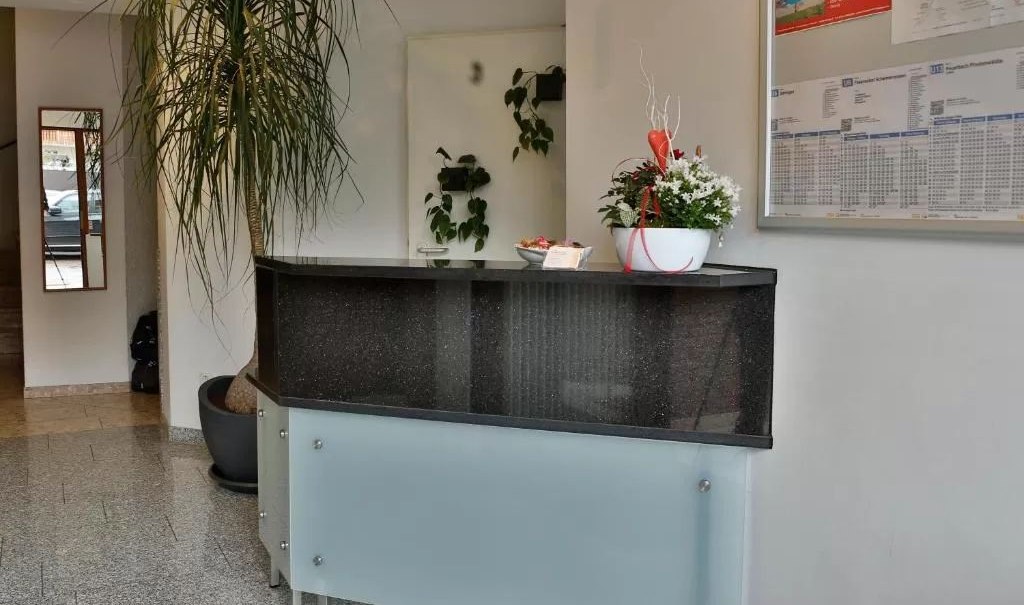 Ein moderner Rezeptionstresen mit einer Pflanze und Dekoration. An der Wand hängt ein Informationsbrett. Der Boden ist gefliest., © Feuerbacher Hof