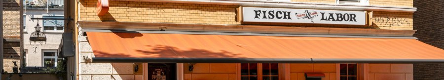 A restaurant called Fischlabor with an orange awning and outdoor seating. The building has yellow brick walls and white window frames., &copy; SMG Stuttgart Marketing GmbH - Sarah Schmid