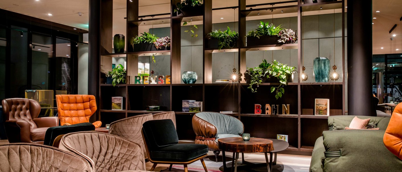 A stylish lobby with comfortable armchairs, plants and decorative shelves. The atmosphere is inviting and modern., &copy; Motel One - Stuttgart Hauptbahnhof