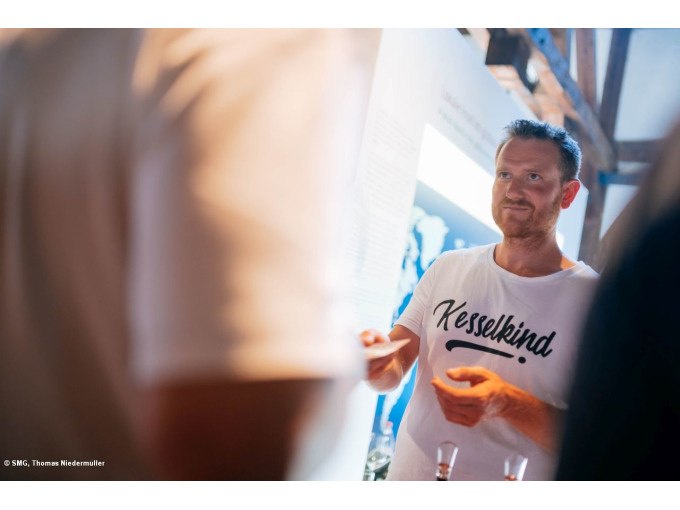 Mann im 'Kesselkinder'-Shirt spricht mit Personen in einem Raum, vermutlich bei einer Weinveranstaltung., © Stuttgart Marketing GmbH Mann im 'Kesselkinder'-Shirt spricht mit Personen in einem Raum, vermutlich bei einer Weinveranstaltung., © Stuttgart Marketing GmbH