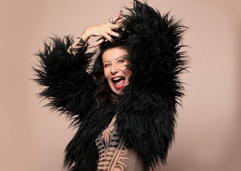 Eine Frau in einem schwarzen, flauschigen Outfit lacht und posiert lebhaft vor einem beigen Hintergrund., &copy; Theaterhaus Stuttgart e.V.