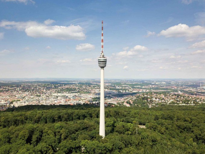 The Stuttgart TV Tower rises above a green forest; the city can be seen in the background, beneath a blue sky with a few clouds., © SWR Media Services GmbH / Fernsehturm Stuttgart The Stuttgart TV Tower rises above a green forest; the city can be seen in the background, beneath a blue sky with a few clouds., © SWR Media Services GmbH / Fernsehturm Stuttgart
