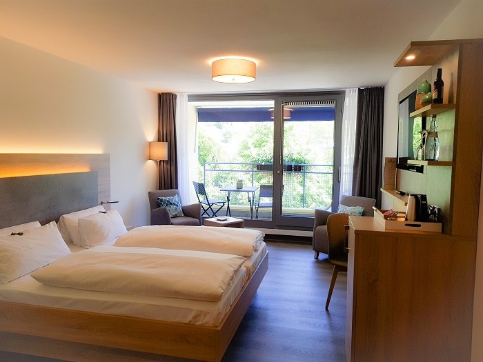 Modernes Hotelzimmer mit Doppelbett, Balkon mit Tisch und Stühlen, gemütlicher Sitzecke und Holzmöbeln. Große Fenster lassen viel Licht herein., © TOMAS