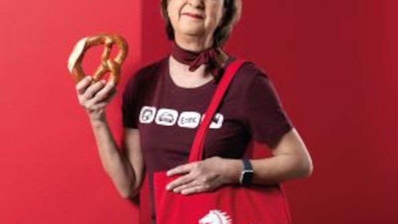 Frau hält eine Brezel und trägt eine rote Tasche mit Pferdemotiv. Sie steht vor einem roten Hintergrund und trägt ein dunkelrotes T-Shirt., © Altes Schauspielhaus und Komödie im Marquardt e.V. Frau hält eine Brezel und trägt eine rote Tasche mit Pferdemotiv. Sie steht vor einem roten Hintergrund und trägt ein dunkelrotes T-Shirt., © Altes Schauspielhaus und Komödie im Marquardt e.V.