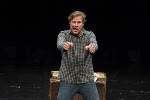 A man in a checked shirt stands on a stage and gesticulates energetically. A dark stage area can be seen in the background., © Theaterhaus Stuttgart e.V. A man in a checked shirt stands on a stage and gesticulates energetically. A dark stage area can be seen in the background., © Theaterhaus Stuttgart e.V.