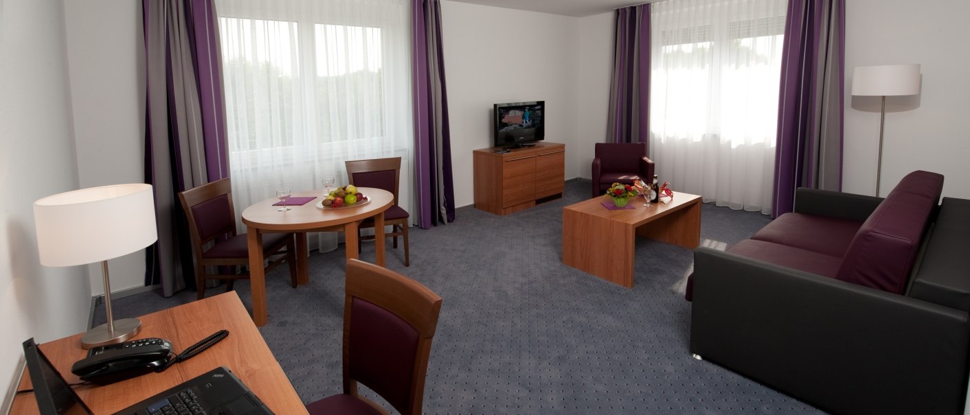 Moderner Wohnbereich einer Suite mit lila Akzenten, Sofa, Tisch, Stühlen, Fernseher und Laptop auf einem Schreibtisch., © Hotel Fortuna