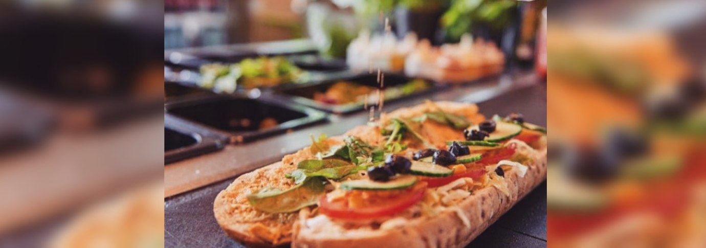 Ein Sandwich wird mit frischem Gem&uuml;se und Oliven belegt. Eine Hand streut Gew&uuml;rze dar&uuml;ber. Im Hintergrund sind Zutaten in Beh&auml;ltern zu sehen., &copy; FreshSub