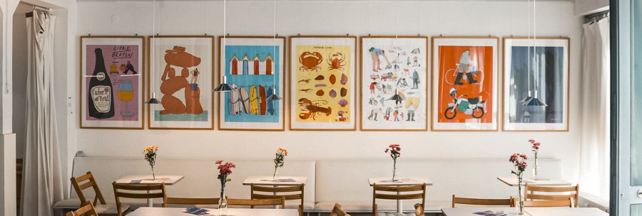 A cosy café with wooden furniture, colorful posters on the wall and flowers on the tables. A projector hangs from the ceiling., © Merlin, Stuttgart