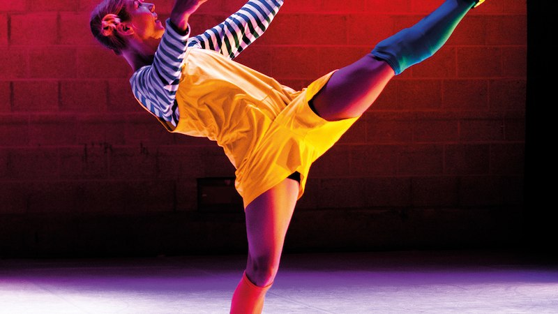 Eine Tänzerin in einem gelben Outfit tanzt auf einer Bühne mit rotem Hintergrund. Sie hebt ein Bein hoch in die Luft., © Theaterhaus Stuttgart e.V.